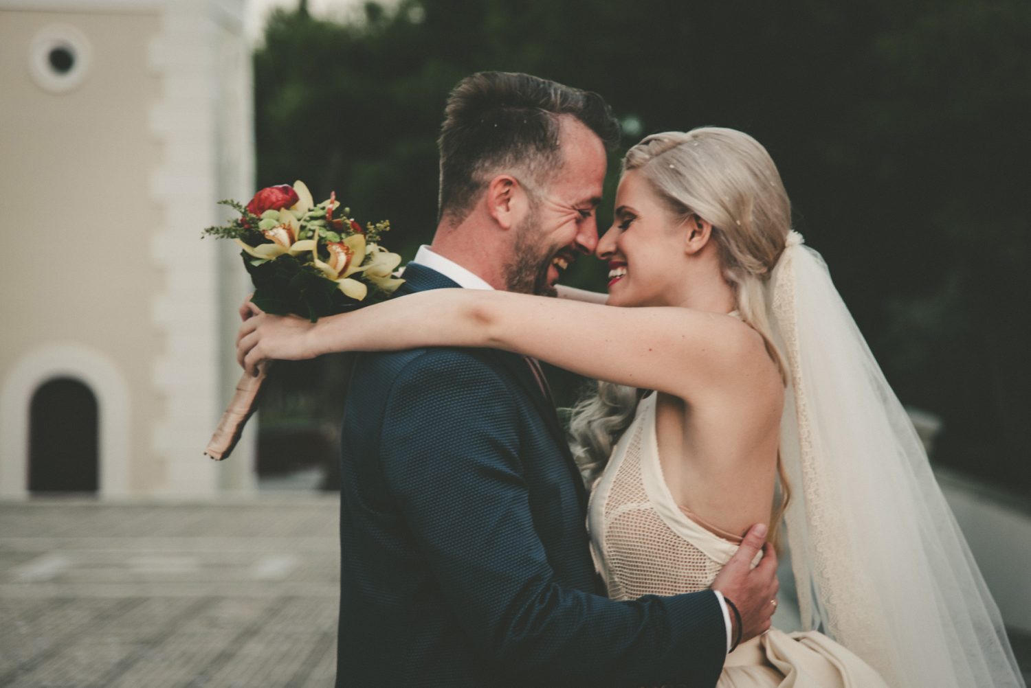 Bride and groom outside the church, about to kiss and holding each other, while Maria wears a back-bow wedding dress by Zolotas with a long veil, and hold a colorful bouquet