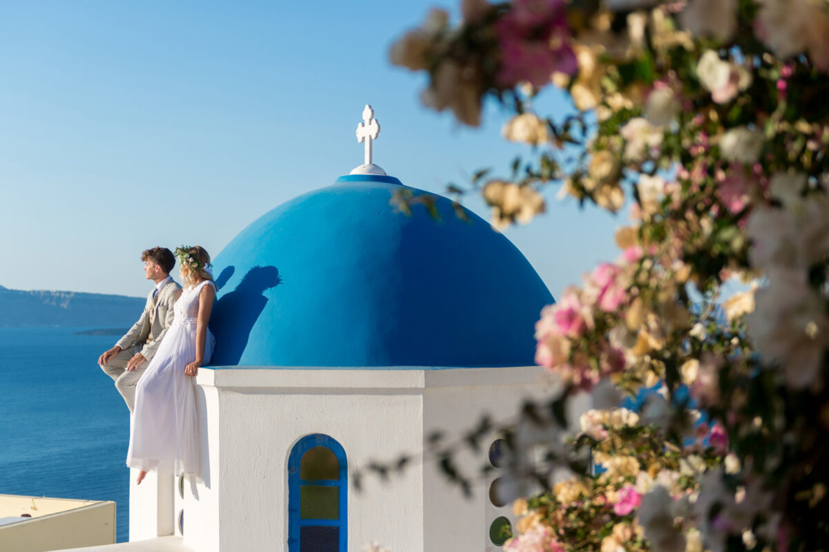 Jess and Stephen in wedding attire, sitting on a Santorini blue dome, framed by bright bougainvilleas: a moment from the Flying the Nest day-after shoot